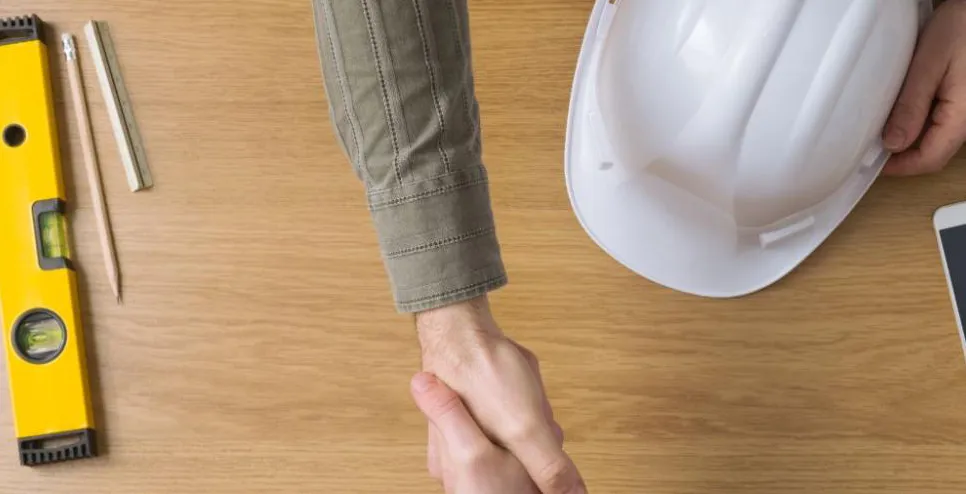 Image of hands shaking across a desk. Level, Architectural Scale Ruler, and pencil on left. Hand on white hard hat next to phone on right.