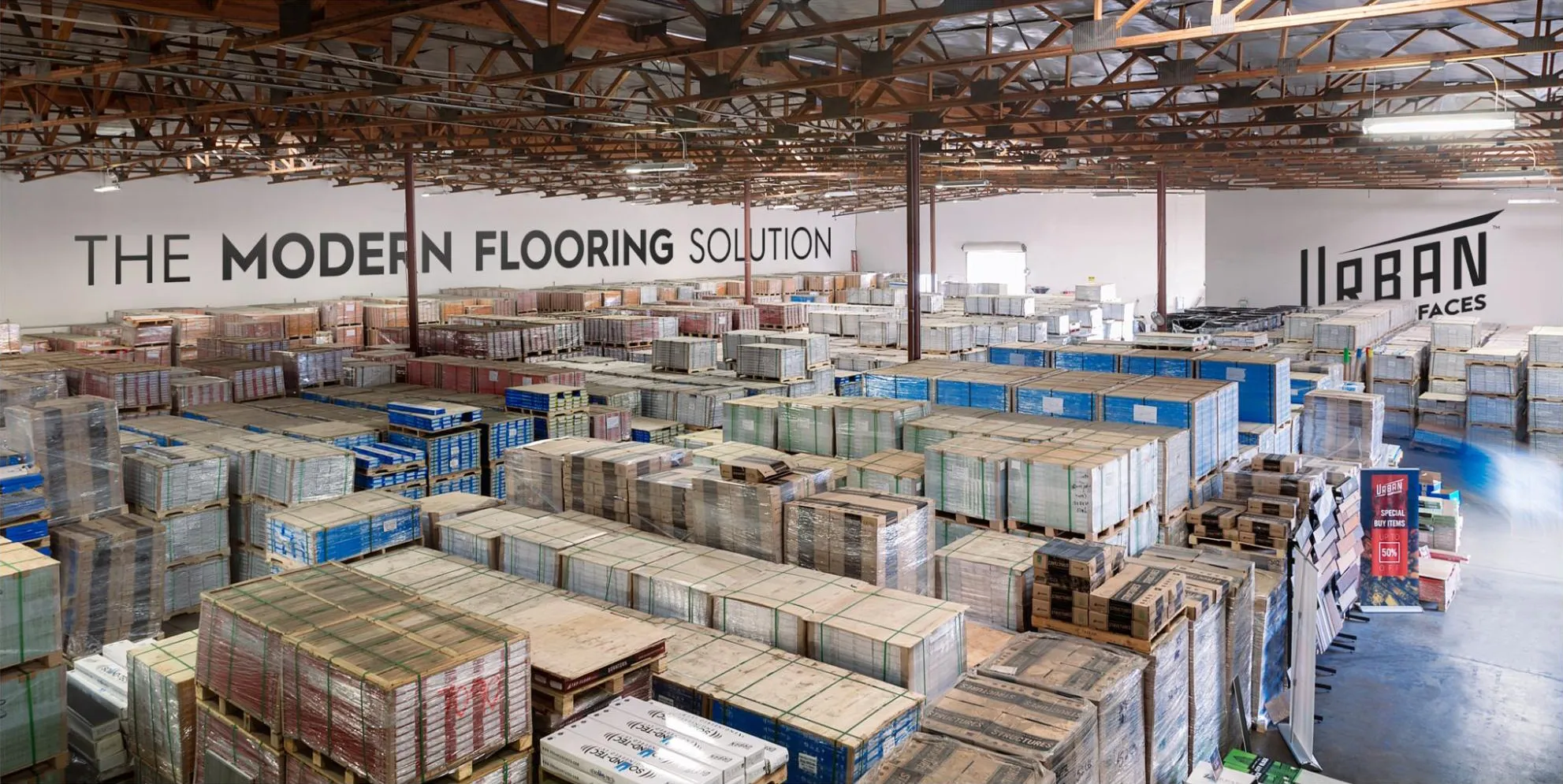 Image of the Urban Surfaces warehouse with pallets of flooring plank boxes. The words "The Modern Flooring Solution" and the Urban Surfaces logo are on the walls.